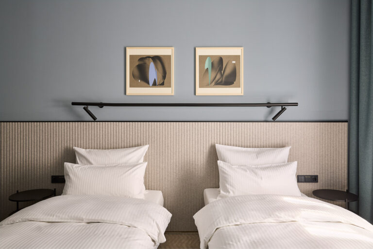 Nowoczesny pokój hotelowy Double Twin Room z dwoma osobnymi łóżkami jednoosobowymi, wspólnym jasnym zagłówkiem oraz dwoma minimalistycznymi obrazami na niebiesko-szarej ścianie.