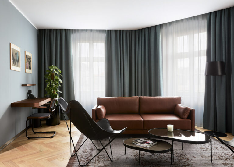 Elegancki salon w apartamencie Executive Suite ze skórzaną brązową kanapą, fotelem motylkowym, kącikiem do pracy z biurkiem oraz dużymi oknami z ciemnymi zasłonami.