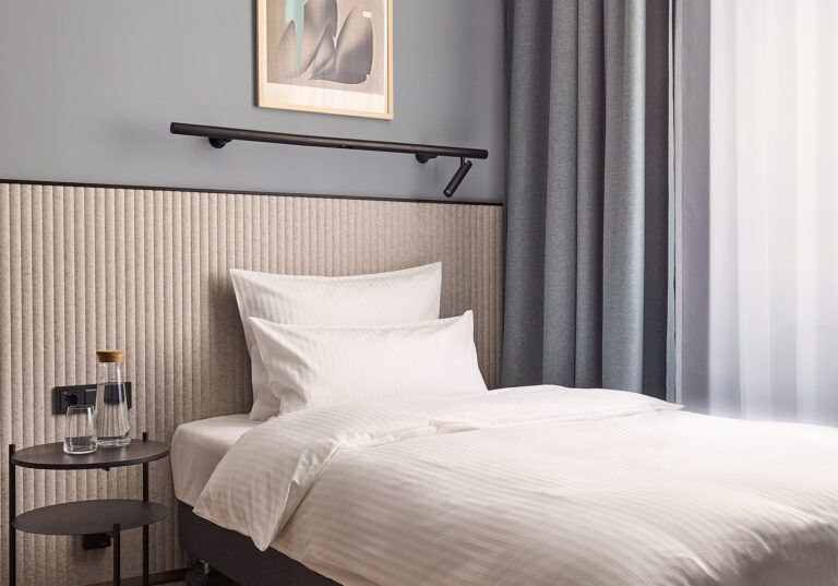 Nowoczesny pokój jednoosobowy Cosy Single Room z pojedynczym łóżkiem kontynentalnym, białą pościelą, tapicerowanym jasnym zagłówkiem i minimalistycznym czarnym stolikiem nocnym.