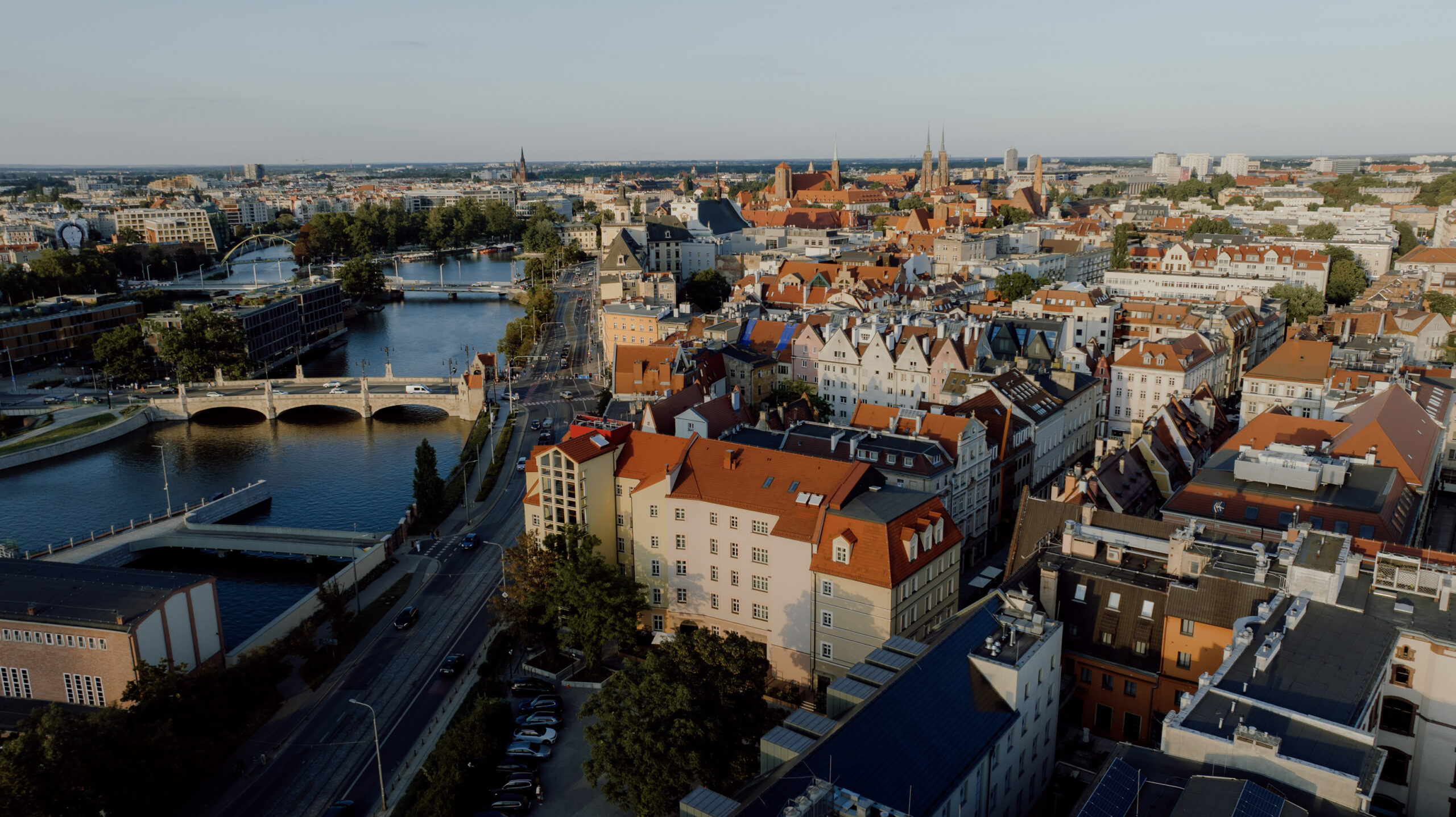 Malownicza panorama Wrocławia z lotu ptaka. Na pierwszym planie widoczna jest elegancka kamienica, w której mieści się Entre Hotel. Po lewej stronie przepływa rzeka Odra spięta historycznymi mostami, a tło stanowi gęsta zabudowa miejska z zabytkowymi wieżami Ostrowa Tumskiego na horyzoncie.