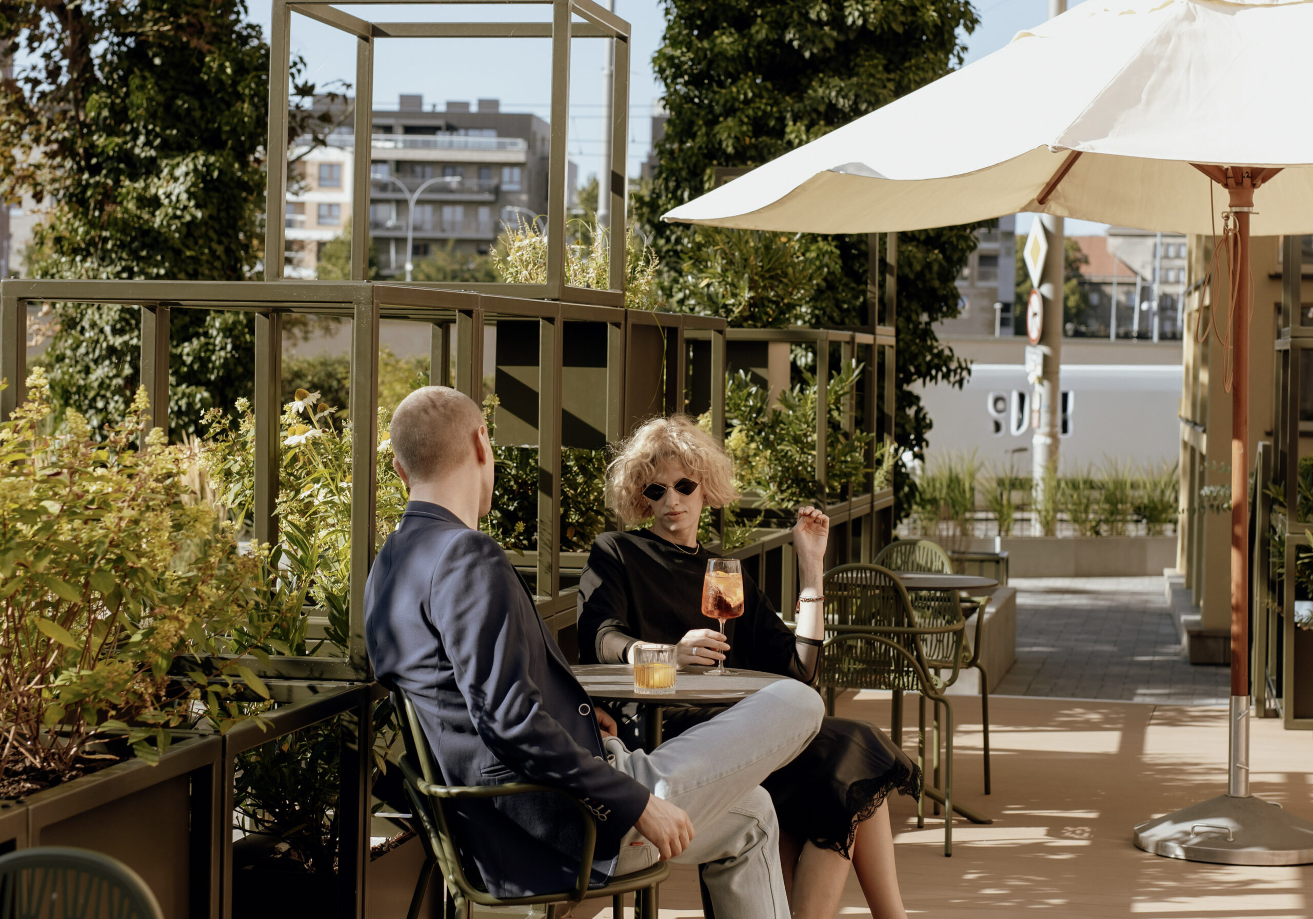 Letni ogródek restauracji Entre we Wrocławiu od strony ulicy Nowy Świat. Para gości odpoczywa przy stoliku pod dużymi parasolami, pijąc koktajle w otoczeniu nowoczesnych, zielonych pergoli z roślinnością.