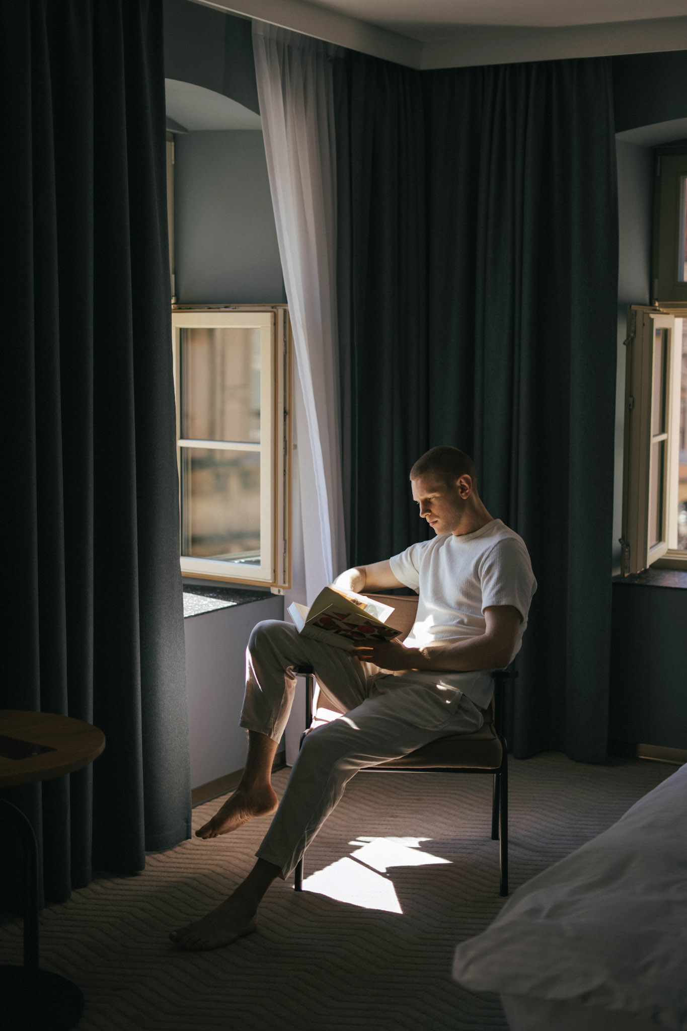 Atmosfera sprzyjająca wyciszeniu w apartamencie. Mężczyzna w białej koszulce siedzi w fotelu i czyta książkę w strumieniu naturalnego światła wpadającego przez duże, otwarte okno. Wnętrze otaczają ciężkie, ciemne zasłony w głębokim odcieniu zieleni lub grafitu, tworząc intymny i spokojny nastrój.