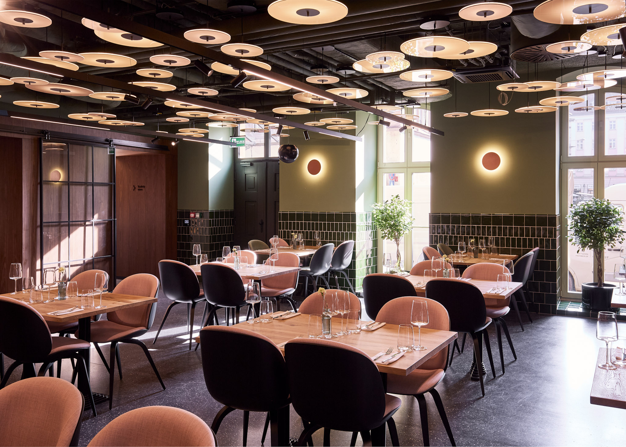 Nowoczesne wnętrze restauracji Entre w stylu brasserie z drewnianymi stolikami, pastelowo-różowymi i czarnymi krzesłami oraz unikalnym sufitem pokrytym okrągłymi lampami.