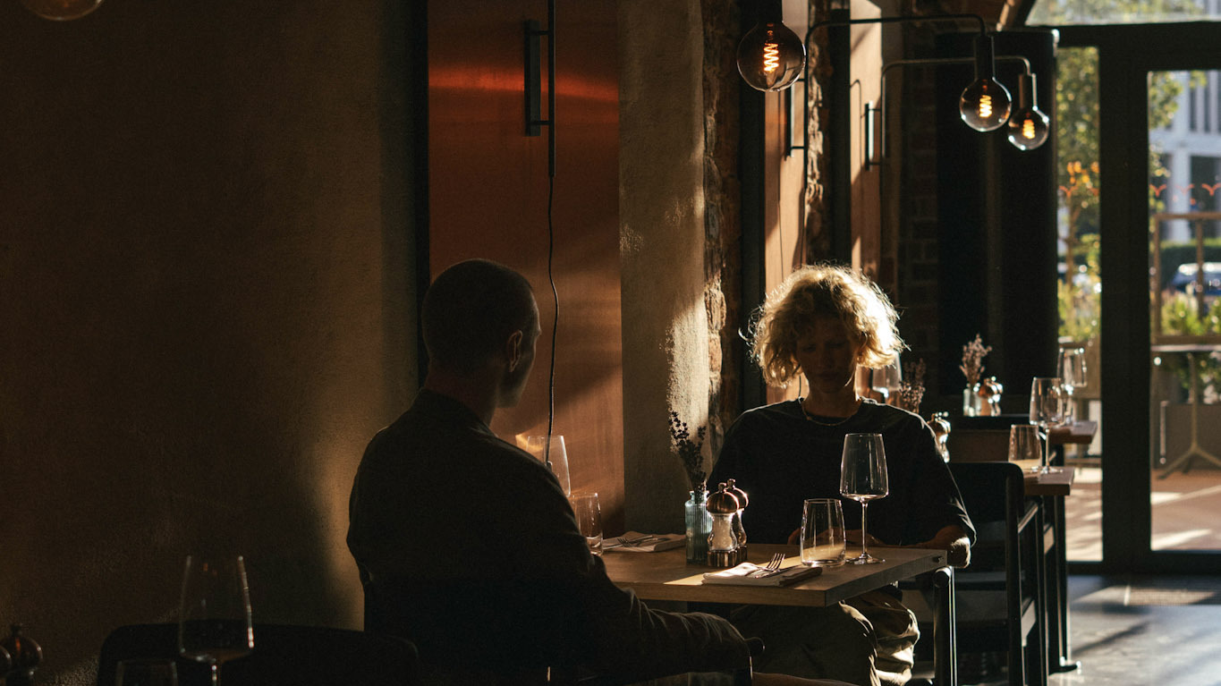 Mężczyzna i kobieta siedzą naprzeciwko siebie przy drewnianym stoliku restauracyjnym. Wnętrze jest urządzone w stylu industrialnym z ciepłym, nastrojowym światłem ozdobnych żarówek, z kieliszkami do wina na stole i dużymi oknami wychodzącymi na zewnątrz.
