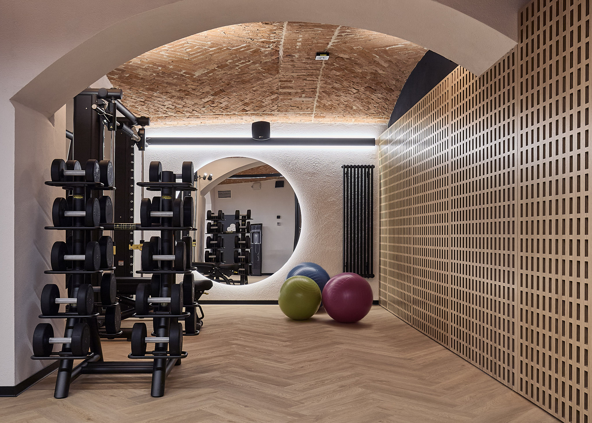 Nowoczesna hotelowa sala fitness ze stojakiem na hantle, piłkami gimnastycznymi i dużym okrągłym lustrem, umieszczona w klimatycznym wnętrzu z ceglanym sklepieniem łukowym.