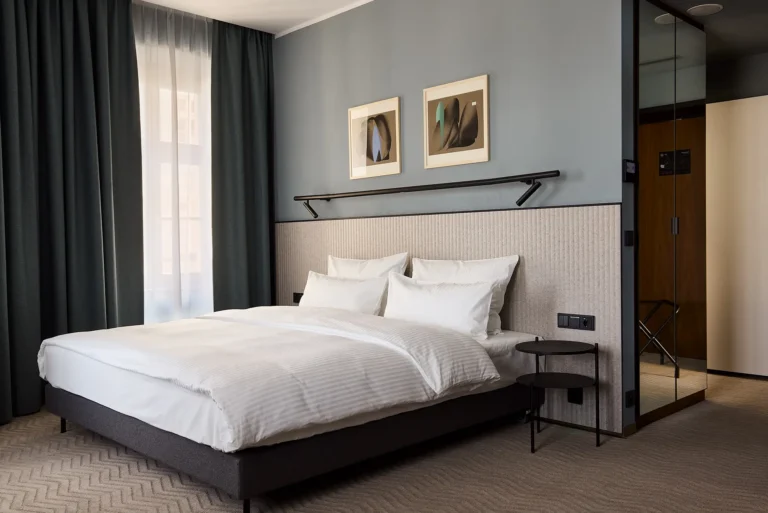 Luksusowa, nowoczesna sypialnia w apartamencie Executive Suite. Elegancki i minimalistyczny wystrój z dużym łóżkiem, białą pościelą, teksturowanym zagłówkiem i ciemnymi zasłonami.