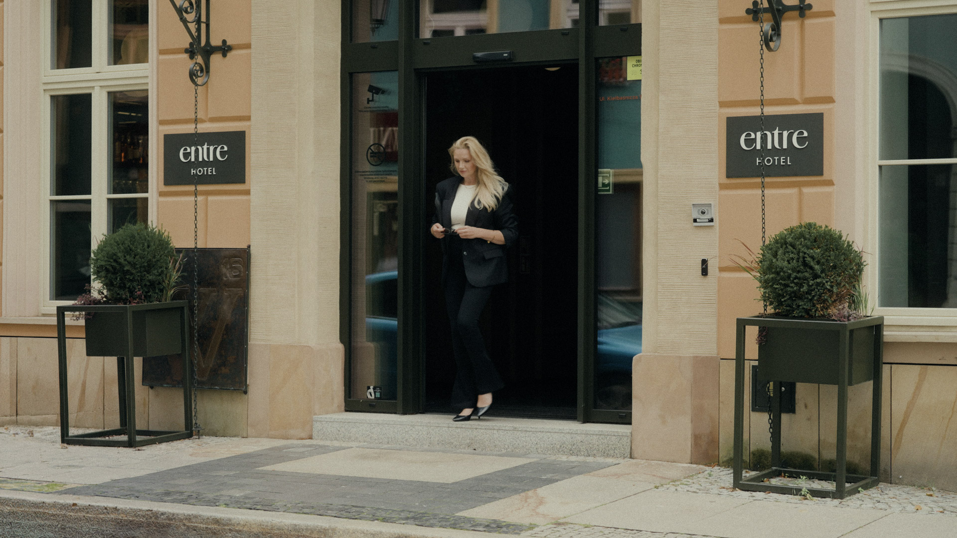 Widok na frontowe wejście do Entre Hotel we Wrocławiu. Blondynka w biznesowym stroju przechodzi przez automatyczne drzwi. Po bokach widoczne czarne tablice z logo hotelu oraz nowoczesne, geometryczne donice z zielonymi krzewami.